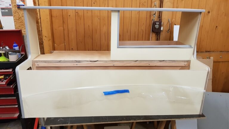 Une armoire en bois partiellement assemblée avec une finition crème clair, placée dans un atelier. Le panneau arrière est incomplet, montrant du bois exposé. Un morceau de ruban adhésif bleu est visible sur le panneau avant. Les outils et létabli environnants sont partiellement visibles.