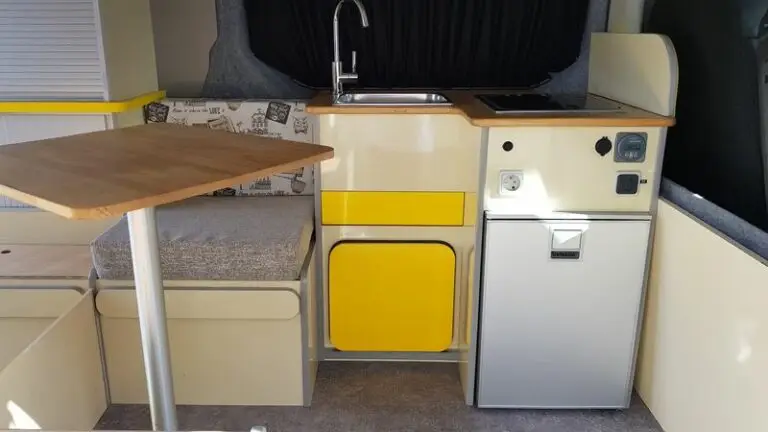 Intérieur d'un Renault Trafic L2H1 comprenant une cuisine compacte avec un petit évier, un feu gaz et un mini-réfrigérateur 12v. On y trouve une table en bois fixée au sol, Des sièges avec coussins, des coffres et un tiroir coulissant.